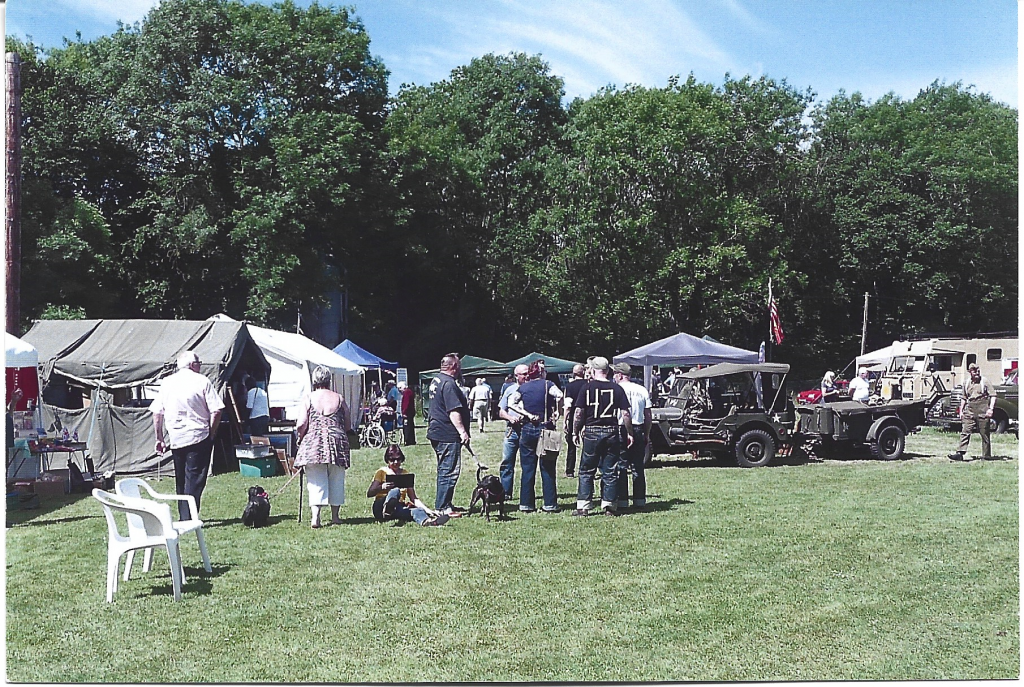 Gazebos and old war vehicles in field with people milling about